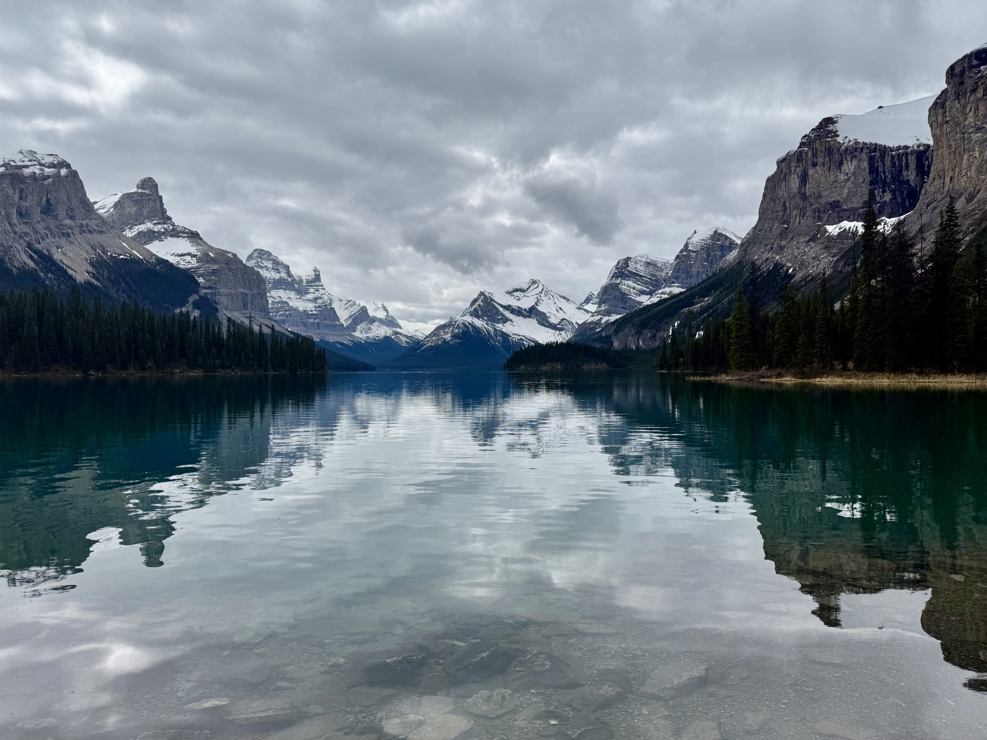 Still glacial water between snow-topped peaks, forest along the shoreline